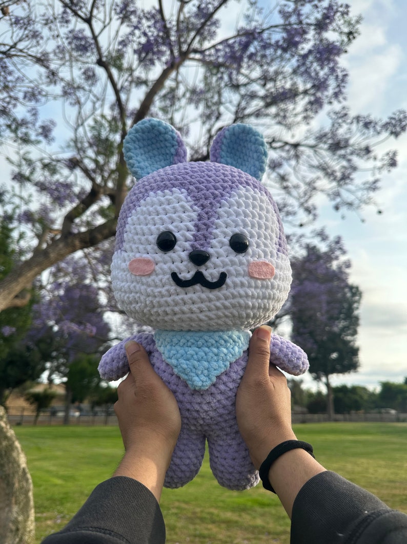 Puede incluir: Un juguete de peluche de crochet con forma de conejo. El conejo es morado y blanco con orejas azules y una bufanda azul. El conejo est&aacute; siendo sostenido por dos manos.