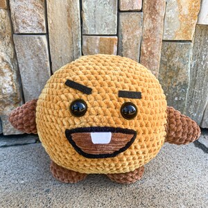 May include: A brown crocheted plush toy with a cartoon face. The toy has black eyebrows, large black eyes, a brown mouth, and a single white tooth.