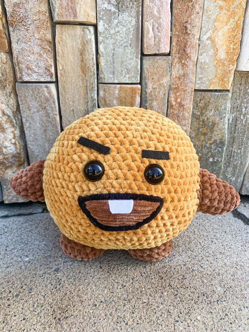 May include: A brown crocheted plush toy with a cartoon face. The toy has black eyebrows, large black eyes, a brown mouth, and a single white tooth.