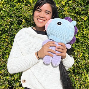 May include: A person is holding a light blue and purple crocheted plush toy with a large black eye. The toy has a pink nose and purple ears. The person is wearing a white sweater and black pants.