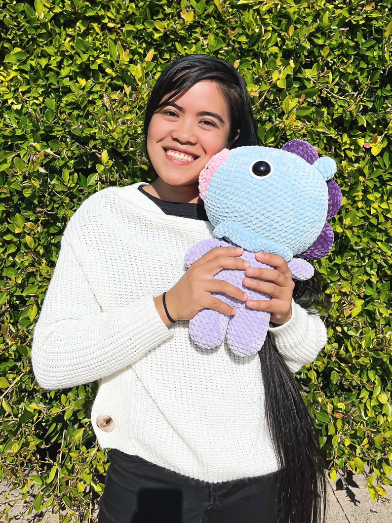 May include: A person is holding a light blue and purple crocheted plush toy with a large black eye. The toy has a pink nose and purple ears. The person is wearing a white sweater and black pants.
