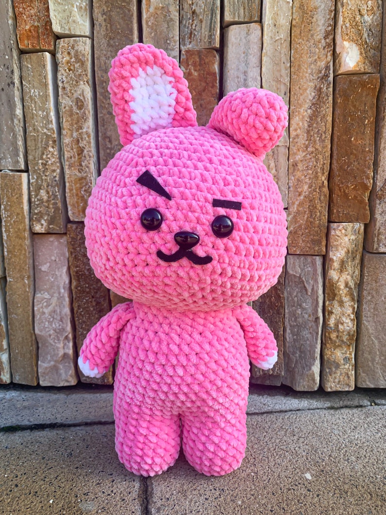 May include: Pink crocheted plush toy shaped like a rabbit with white ears and black eyes. The toy has a grumpy expression with a black line above each eye.