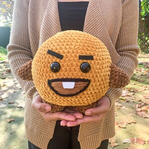 May include: A crocheted plush toy in the shape of a brown bear with a grumpy expression. The bear has black eyes, a black mouth, and a white tooth. The bear is being held by a person wearing a brown cardigan.