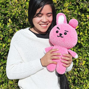 May include: A person is holding a pink crocheted bunny plush toy with white ears and black eyes. The bunny has a frowning expression.