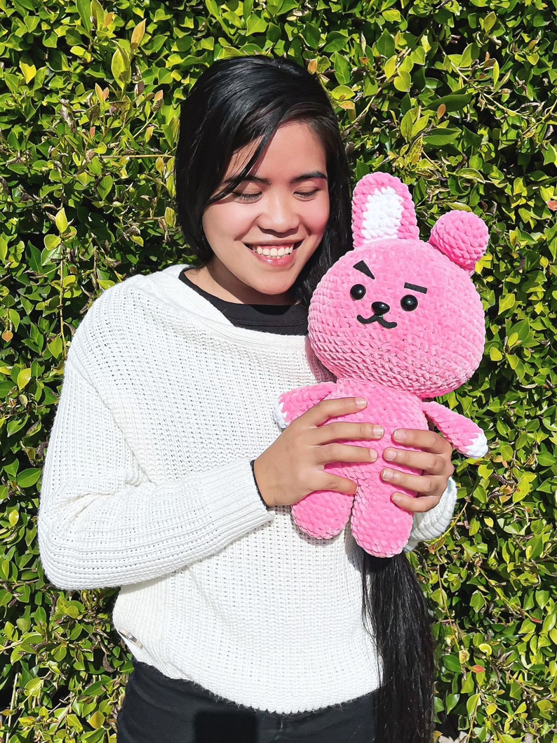 May include: A person is holding a pink crocheted bunny plush toy with white ears and black eyes. The bunny has a frowning expression.