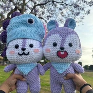 Puede incluir: Dos peluches de crochet, uno con un gorro de dinosaurio azul y morado, el otro con orejas de conejo. Ambos son morados y blancos con caras bordadas y bufandas azules.