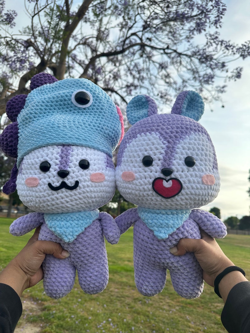 Puede incluir: Dos peluches de crochet, uno con un gorro de dinosaurio azul y morado, el otro con orejas de conejo. Ambos son morados y blancos con caras bordadas y bufandas azules.