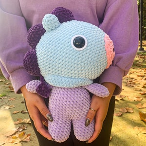 May include: A crocheted plush toy in the shape of a purple and blue creature with a single large eye. The toy is being held by a person wearing a purple sweater and black pants.