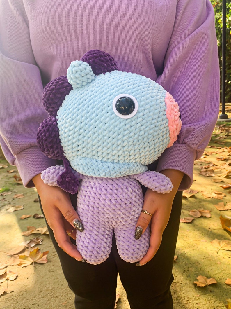May include: A crocheted plush toy in the shape of a purple and blue creature with a single large eye. The toy is being held by a person wearing a purple sweater and black pants.