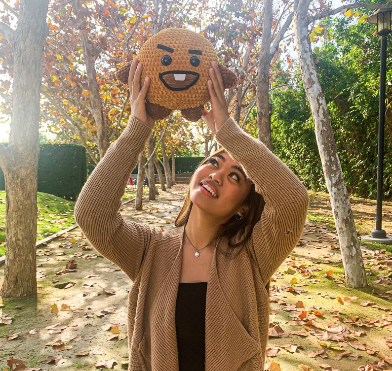 May include: A person wearing a brown cardigan holds a brown plush toy with a cartoon face. The toy has a brown body with a white face and black eyes. The person is standing in a park with trees and leaves on the ground.