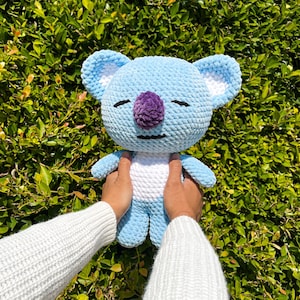 May include: A light blue crocheted koala plush toy with a white belly and a purple nose. The toy is being held by a person's hands.