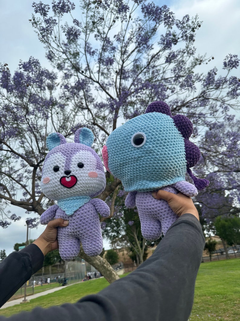 Puede incluir: Dos peluches de crochet, uno morado con cara de conejo y el otro azul con cara de pez, se sostienen en alto frente a un &aacute;rbol con flores moradas.