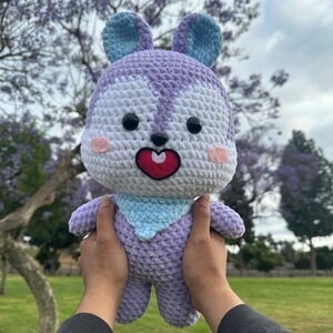 Puede incluir: Un juguete de peluche tejido a crochet en forma de conejo morado y blanco con orejas azules. El conejo tiene una nariz roja en forma de coraz&oacute;n y ojos negros. El conejo est&aacute; siendo sostenido por las manos de una persona.