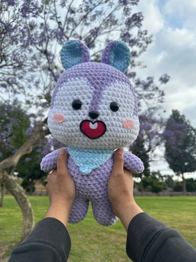 Puede incluir: Un juguete de peluche tejido a crochet en forma de conejo morado y blanco con orejas azules. El conejo tiene una nariz roja en forma de coraz&oacute;n y ojos negros. El conejo est&aacute; siendo sostenido por las manos de una persona.