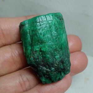 May include: A raw, cylindrical emerald crystal with a deep green color and a textured, uneven surface. The stone features vertical striations and natural fissures throughout its structure, displaying a matte finish with varying shades of green.