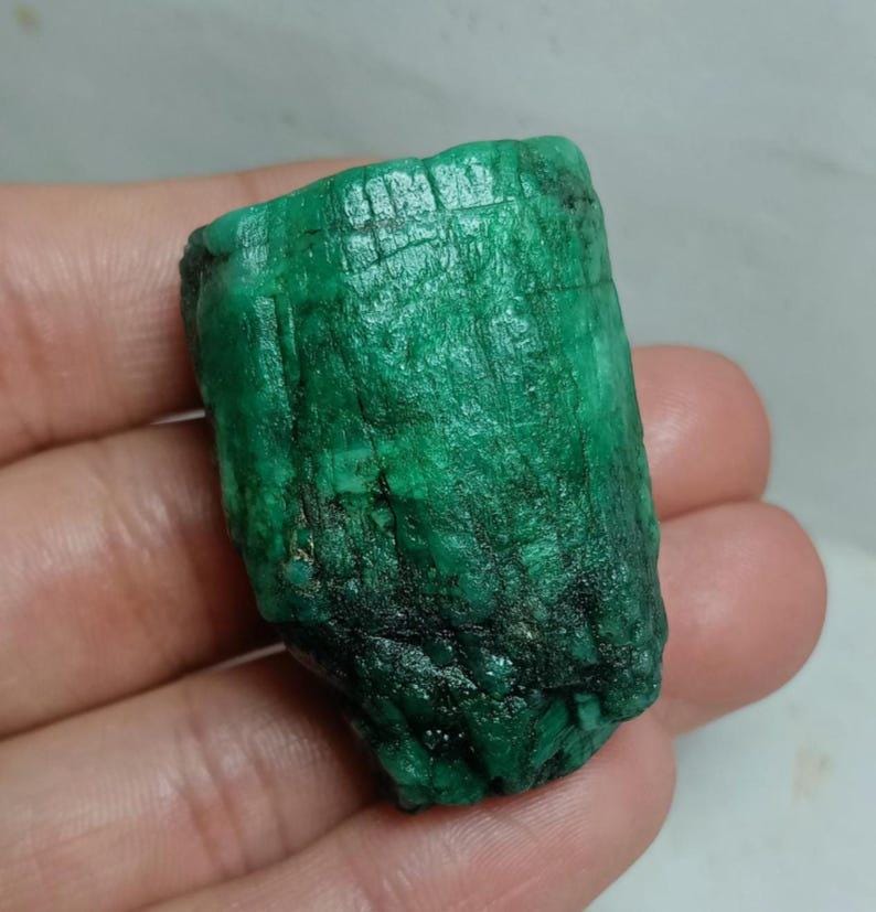 May include: A raw, cylindrical emerald crystal with a deep green color and a textured, uneven surface. The stone features vertical striations and natural fissures throughout its structure, displaying a matte finish with varying shades of green.