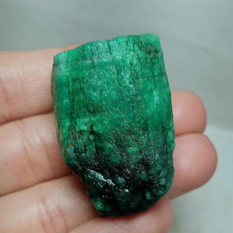 May include: A raw, uncut emerald crystal with a deep green color and a textured, columnar surface. The stone features vertical striations and natural variations in opacity, displaying a mix of vibrant green hues and darker, opaque mineral inclusions.