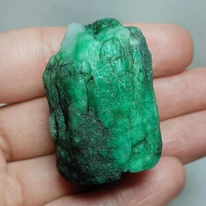 May include: A raw, uncut emerald gemstone with a deep green color and a textured, crystalline surface. The stone features natural variations in opacity and tone, with a prominent white inclusion visible at the top edge of the mineral specimen.