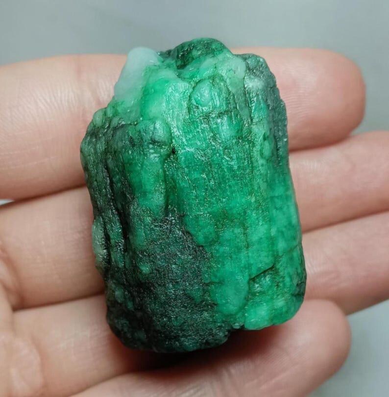 May include: A raw, uncut emerald gemstone with a deep green color and a textured, crystalline surface. The stone features natural variations in opacity and tone, with a prominent white inclusion visible at the top edge of the mineral specimen.