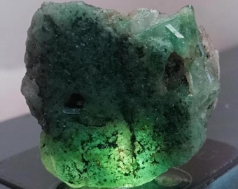 May include: A raw, unpolished emerald crystal specimen featuring a vibrant green hue with translucent sections and dark mineral inclusions. The stone displays a rough, jagged texture with natural crystalline formations and varying levels of opacity.