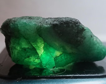 May include: A raw, uncut emerald gemstone displaying a deep forest green color with translucent sections that allow light to pass through. The stone has a rough, irregular surface texture with visible crystalline structures and dark inclusions.