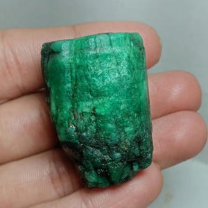 May include: A raw, cylindrical emerald crystal with a vibrant green hue and a textured, uneven surface. The stone displays natural vertical striations and a semi-opaque appearance, showcasing its unpolished, geological form in a natural state.