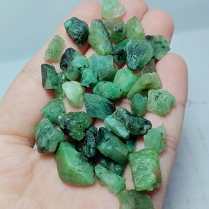 May include: A collection of rough, green emerald gemstones in various sizes and shapes. The emeralds are scattered in a hand, showcasing their natural beauty and raw form.