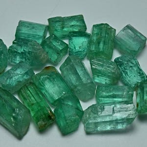 Könnte beinhalten: Eine Ansammlung roher, ungeschliffener Smaragdkristalle in verschiedenen leuchtenden Grüntönen. Diese natürlichen Edelsteine weisen hexagonale prismatische Strukturen mit sichtbaren Einschlüssen, unterschiedlicher Transparenz und rauen, gezackten Kanten auf.
