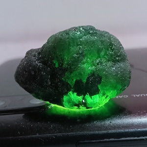 May include: A large, dark green, rough-cut gemstone with a bright green glow. The gemstone is sitting on a black surface.