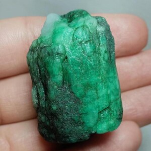 May include: A raw, uncut emerald gemstone with a deep green color and a rough, textured surface. The stone features natural variations in opacity and tone, with a small patch of lighter, translucent white material visible at the top edge.