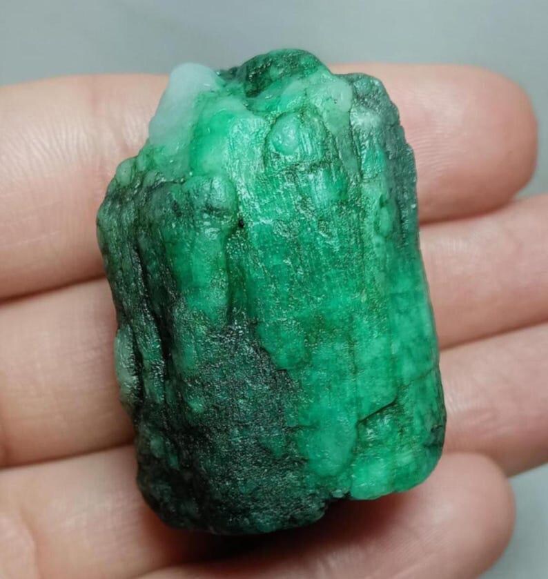 May include: A raw, uncut emerald gemstone with a deep green color and a rough, textured surface. The stone features natural variations in opacity and tone, with a small patch of lighter, translucent white material visible at the top edge.