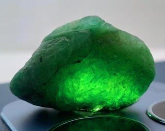 May include: A large, green, translucent gemstone with a rough, irregular shape. The stone is illuminated from below, highlighting its internal structure.