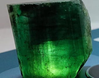 May include: A raw, translucent emerald crystal with a deep green hue and visible internal fractures. The stone features a blocky, prismatic shape with natural vertical striations along its surface, displaying varying levels of light transparency.