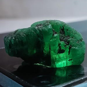 May include: A raw, uncut emerald gemstone with a deep green hue and a rough, textured surface. The gemstone has dark inclusions and a natural, organic shape, reflecting light and displaying a vibrant color.