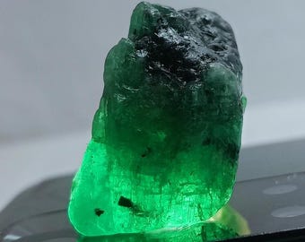 May include: A large, rough, green emerald gemstone with black inclusions. The emerald is translucent and has a glassy luster.