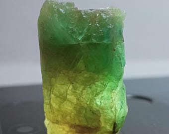 May include: A raw, natural gemstone with a gradient of colors. The top is a vibrant green, transitioning to a lighter green and then a yellow-green at the base. The stone has a rough, textured surface.