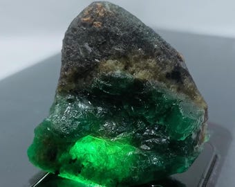 May include: A large, rough, green emerald gemstone with black and brown inclusions. The emerald is illuminated from below, highlighting its green color.