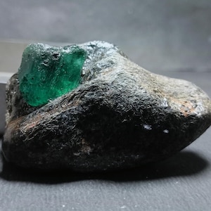 May include: A raw, vibrant green emerald crystal embedded within a dark, textured, and irregular rock matrix. The gemstone displays a translucent quality and a jagged, natural shape, contrasting sharply against the rough, dark gray stone surface.