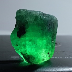 May include: A large, rough, green emerald gemstone with black inclusions. The emerald is sitting on a black surface.