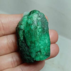 May include: A raw, uncut emerald gemstone with a deep green color and a textured, crystalline surface. The stone features natural variations in opacity and tone, displaying a rugged, unpolished appearance with visible vertical striations.