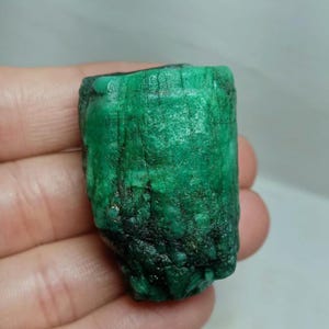 May include: A raw, cylindrical emerald crystal featuring a vibrant, deep green color with a textured, opaque surface. The stone displays natural vertical striations and irregular mineral inclusions throughout its structure, showcasing a rough, unpolished finish.