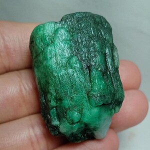 May include: A raw, uncut emerald gemstone with a deep green color and a textured, columnar crystalline structure. The stone displays natural vertical striations and variations in opacity, ranging from translucent to opaque green throughout.