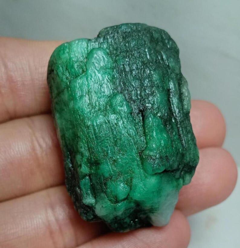 May include: A raw, uncut emerald gemstone with a deep green color and a textured, columnar crystalline structure. The stone displays natural vertical striations and variations in opacity, ranging from translucent to opaque green throughout.