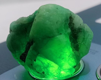 May include: A large, green emerald gemstone with a rough, natural surface. The emerald is illuminated from below, highlighting its translucent quality.