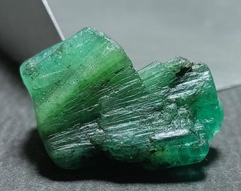 May include: A raw, unpolished emerald crystal with a vibrant green hue and a distinct hexagonal columnar structure. The surface displays natural vertical striations, jagged edges, and varying levels of translucency throughout the stone.