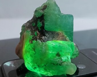 May include: A large, rough, green emerald crystal with a dark brown matrix. The crystal is illuminated from below, causing it to glow green.
