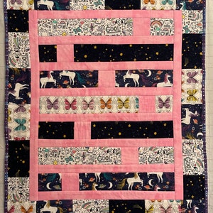 May include: A pink and black patchwork quilt with a unicorn and butterfly theme. The quilt is made up of different fabric squares with various patterns, including stars, rainbows, and doodles. The quilt is edged with a black and white trim.