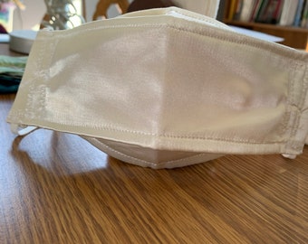 May include: A cream-colored face mask made of a shiny, satin-like fabric. The mask has a pleated design and white elastic ear loops. The stitching is visible along the edges, and the mask appears to be handmade.