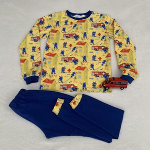 May include: A children's pajama set featuring a firefighter design. The yellow top has a pattern of firefighters, houses, and fire engines. The trousers are blue. A red toy fire engine is also visible.