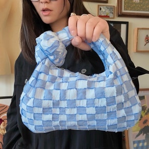 以下が含まれることがあります： 結び目の付いたハンドルが付いた、水色と白のチェック柄のハンドバッグ。バッグは持ち上げられ、織り模様と柔らかな質感を披露しています。バッグのデザインは、繰り返しの正方形のパターンが特徴です。